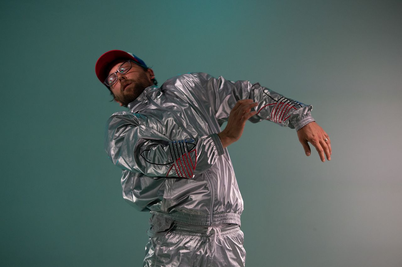 James Gnam, wearing an all-silver outfit and shiny hat, leaning back from the camera with arms raised.