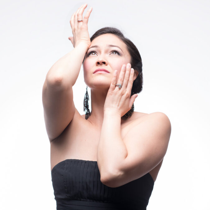 A woman poses with hands touching her face, looking up to the sky. She is wearing a black strapless top and dangling earrings.