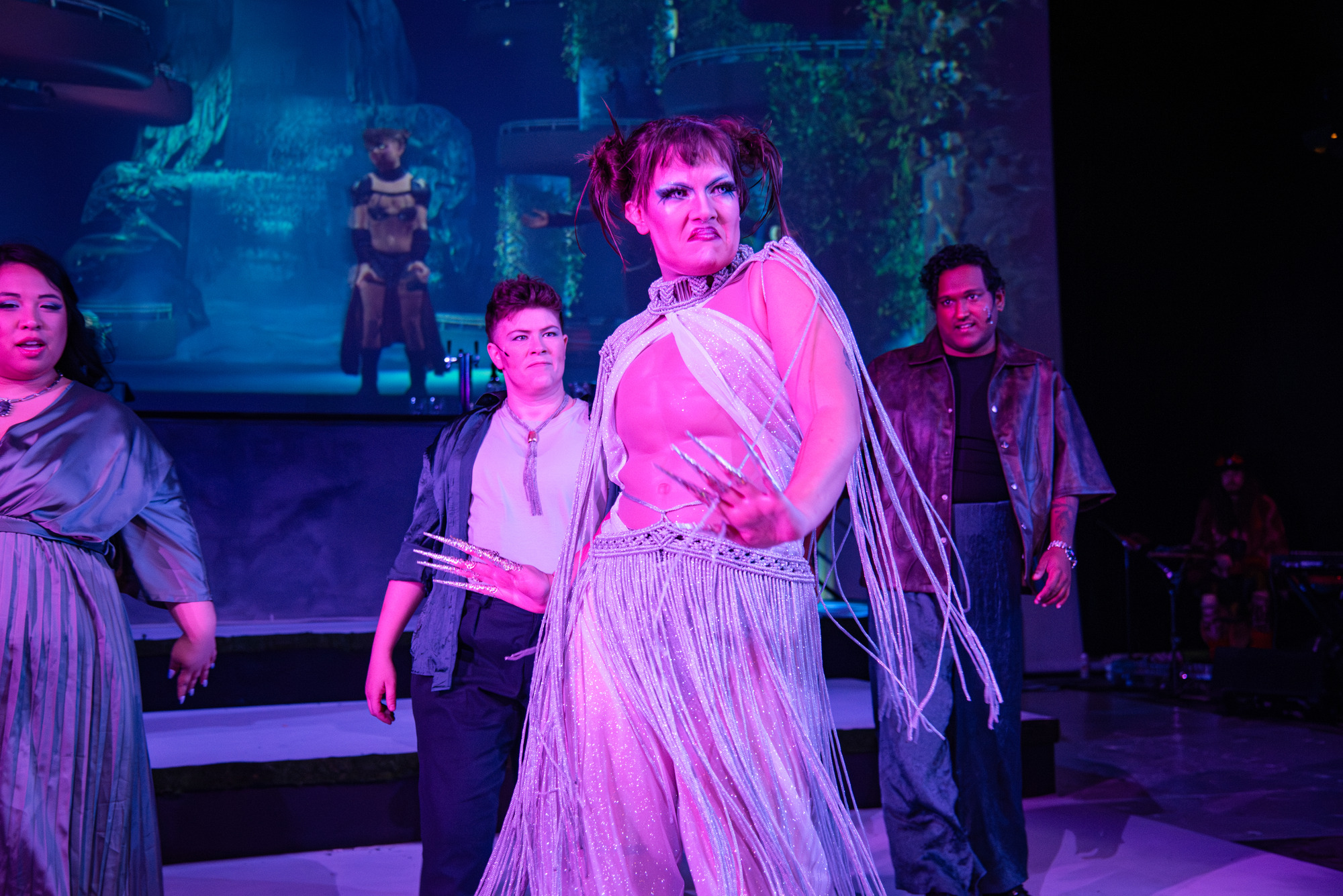 Stage performance with actors bathed in purple and pink light. In the foreground, a performer with long false nails, dramatic makeup and a fringed metallic outfit poses with arms outstretched and a passionate frown. In the background are three other performers wearing  satin outfits, while in the very back a scantily clad person wearing shoulder pads and a cape stands amid a screen of leafy foilage and a castle-like building.