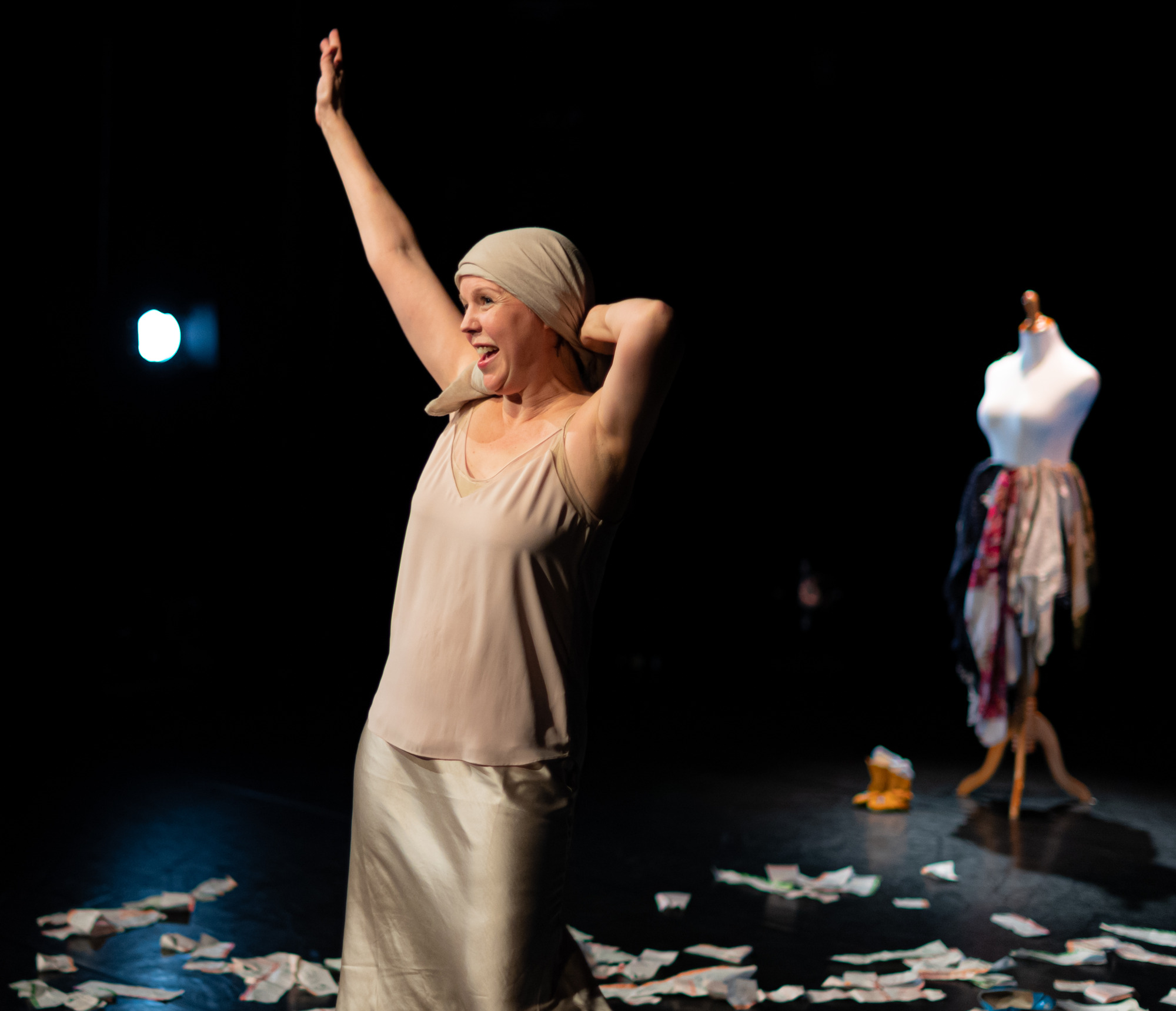 A woman stands on stage, one hand raised and the other clasping a scarf wrapped around her head. She is wearing a neutral-coloured silky tank top and shirt. Onstage there is a half-dressed mannequin and paper scattered on the floor. 