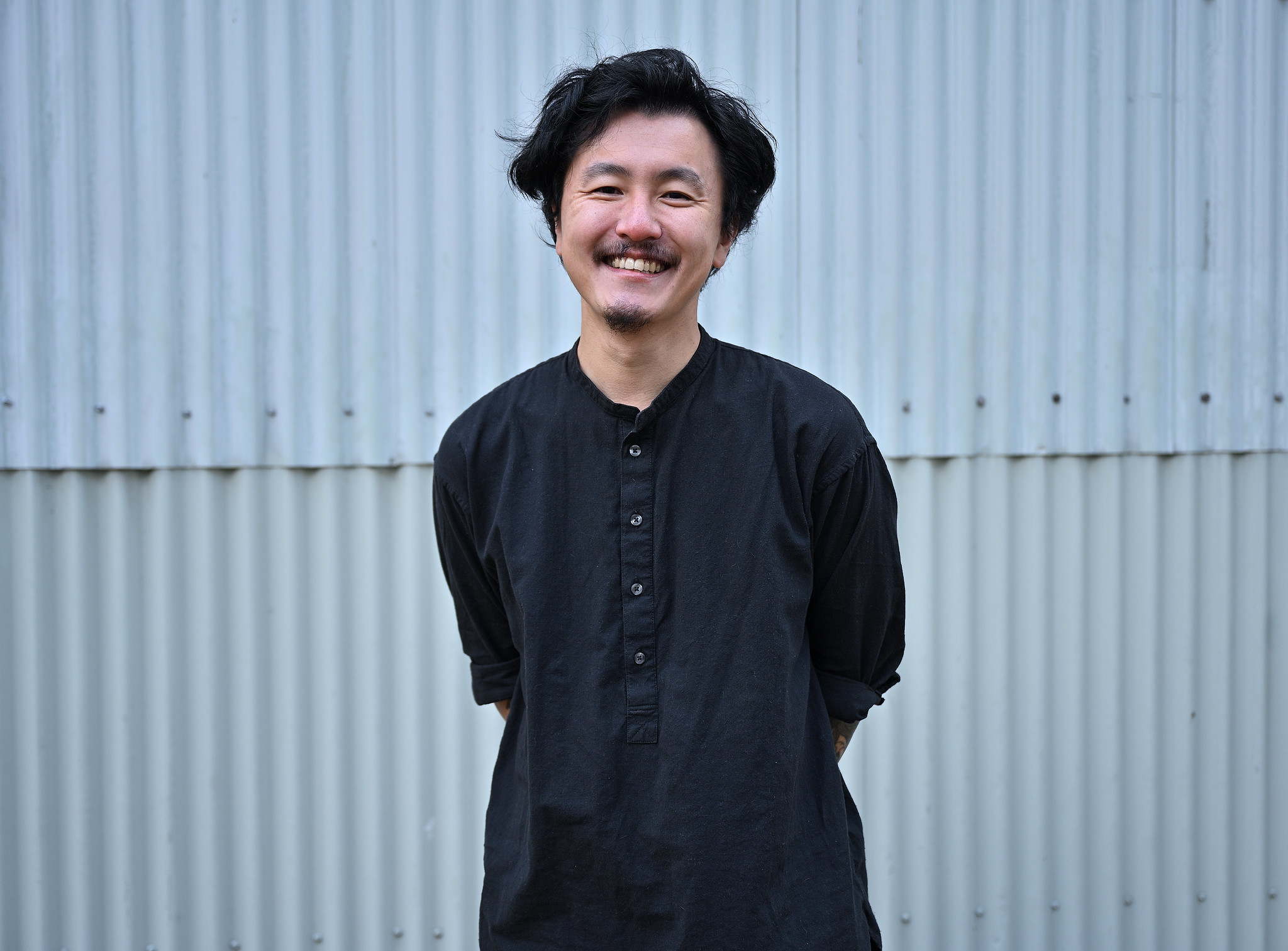 An Asian man smiles at the camera with arms behind his back. He has black floppy hair, a moustache and goatee, and is wearing a collarless black shirt with buttons. The portrait has a backdrop of metal building siding. 