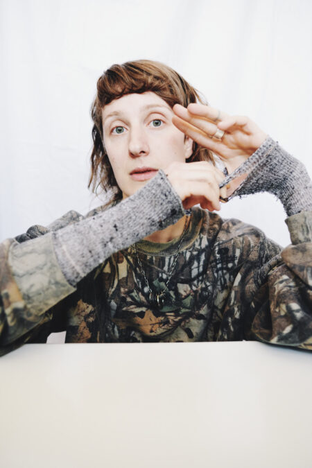 A person poses with arms propped up on a white table, tugging at oustretched fingers and holey sweater cuffs. They have wavy brown hair and short bangs, and are wearing a camouflage sweatshirt and silver chain with ring pendants. They are making eye contact with the camera. 