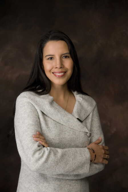 A woman with long dark hair poses smiling in front of a brown backdrop. Her arms are crossed and she wears a light grey wool sweater and silver bracelet.