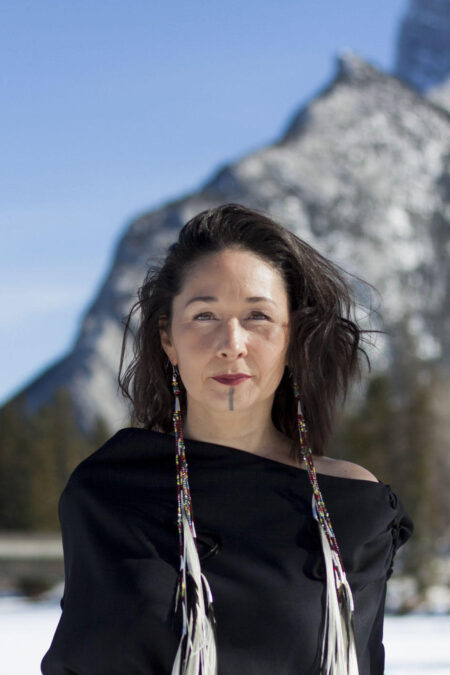 Portrait of a woman standing in front of a winter mountain landscape. She stares directly at the camera, shoulder-length hair blowing in the wind. She has a tattoo from lip to chin, and wears long beaded feather earrings that reach down her torso and a black top. 