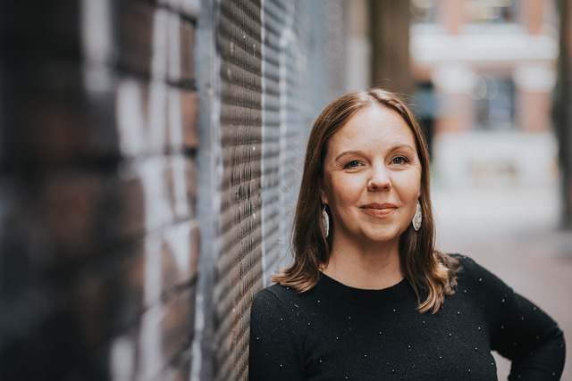 Portrait of a woman with medium-length brown hair and beaded earrings leaning against a painted brick wall.