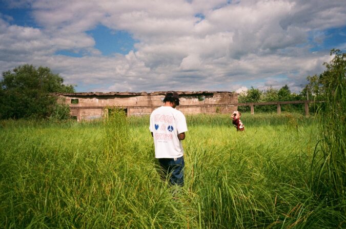 A grassy landscape with cloudy blue sky. In the foreground is a standing person facing away, wearing a white t-shirt that says "Less is more" on the back and blue pants, standing in waist-high grass. In the background is another person wearing a burgundy sports jersey, arms raised. In the far background is a crumbling building.